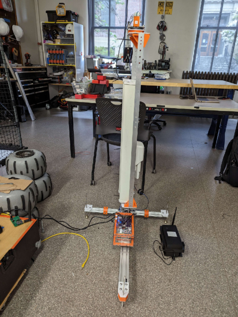 A picture of an antenna system in a crowded lab space. On the floor to the right of the antenna is the black box enclosure that contains the transmitter to send location data from the rover. To the left is the comms station for the rover, and the green receiver board is also visible.